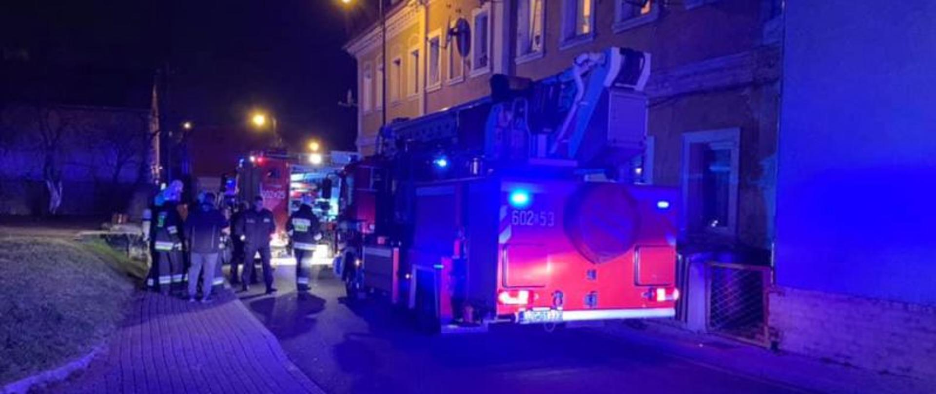 Pora nocna. Na pierwszym planie samochód specjalny podnośnik SHD-25 o numerze operacyjnym 602-53 stoi przed frontem budynku wielorodzinnego. Dalej samochód ratowniczo-gaśniczy. Obok pojazdów stoi grupka strażaków oraz policjantów.