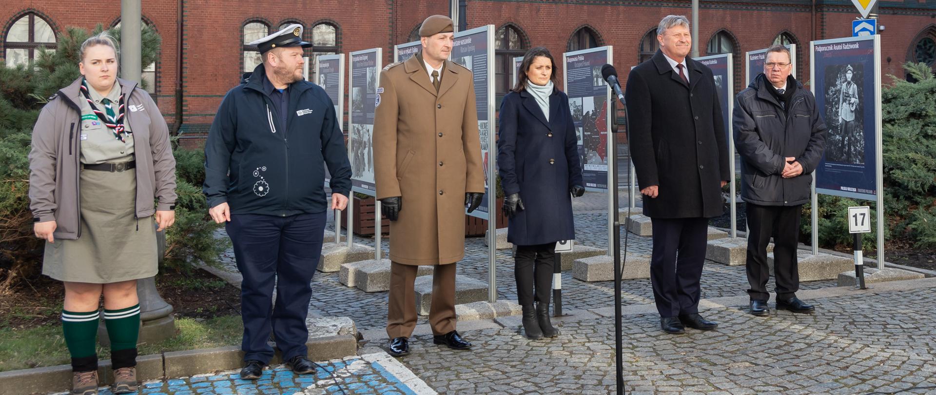 6 osób stojących na tle plansz z wizerunkami żołnierzy AK
