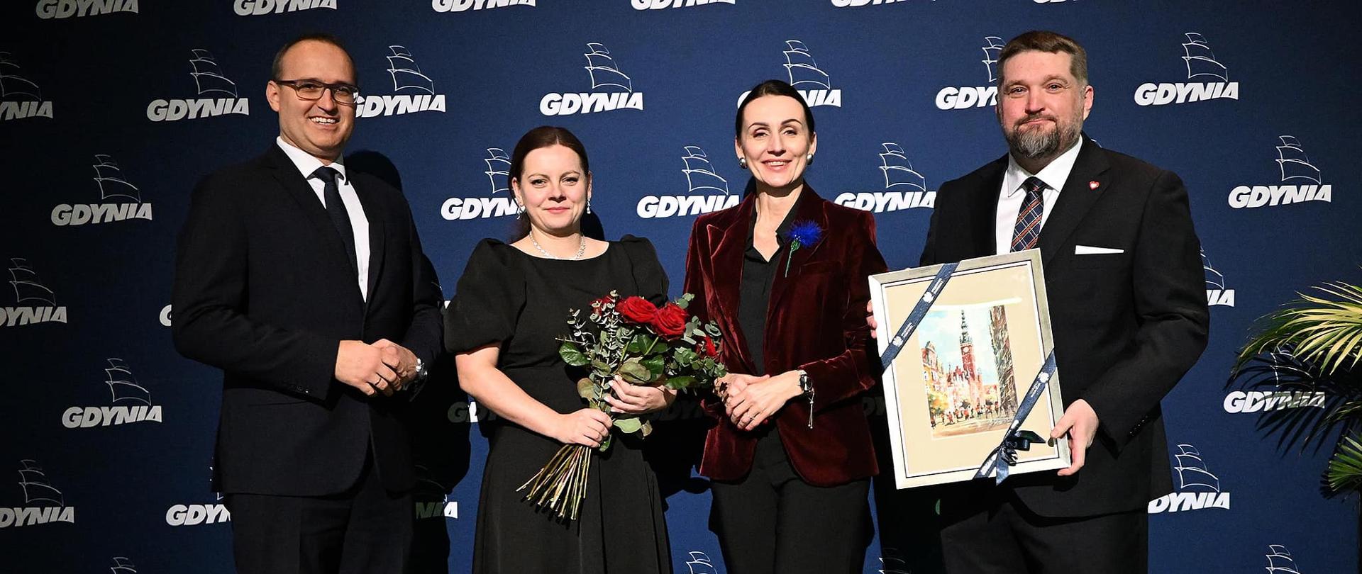 cztery osoby, dwie i kobiety i dwóch mężczyzn stoi na scenie. Mężczyzna trzyma grafikę przedstawiającą stary Gdańsk