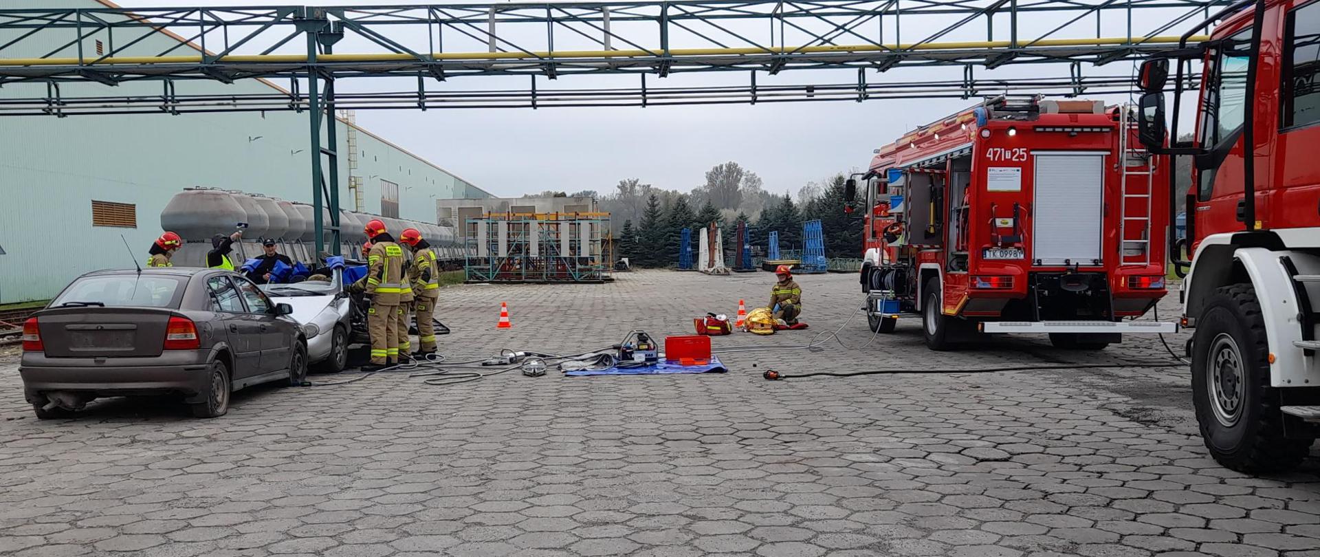 Zdjęcie na terenie zakładu przemysłowego. Po prawej stoją dwa samochody strażackie z których jest wyjęty sprzęt ratowniczy. Obok nich klęczy strażak, który udziela pomocy poszkodowanemu. Po lewej widać dwa samochody osobowe. Przy drugim stoją strażacy, którzy wykonują pracę narzędziami hydraulicznymi.