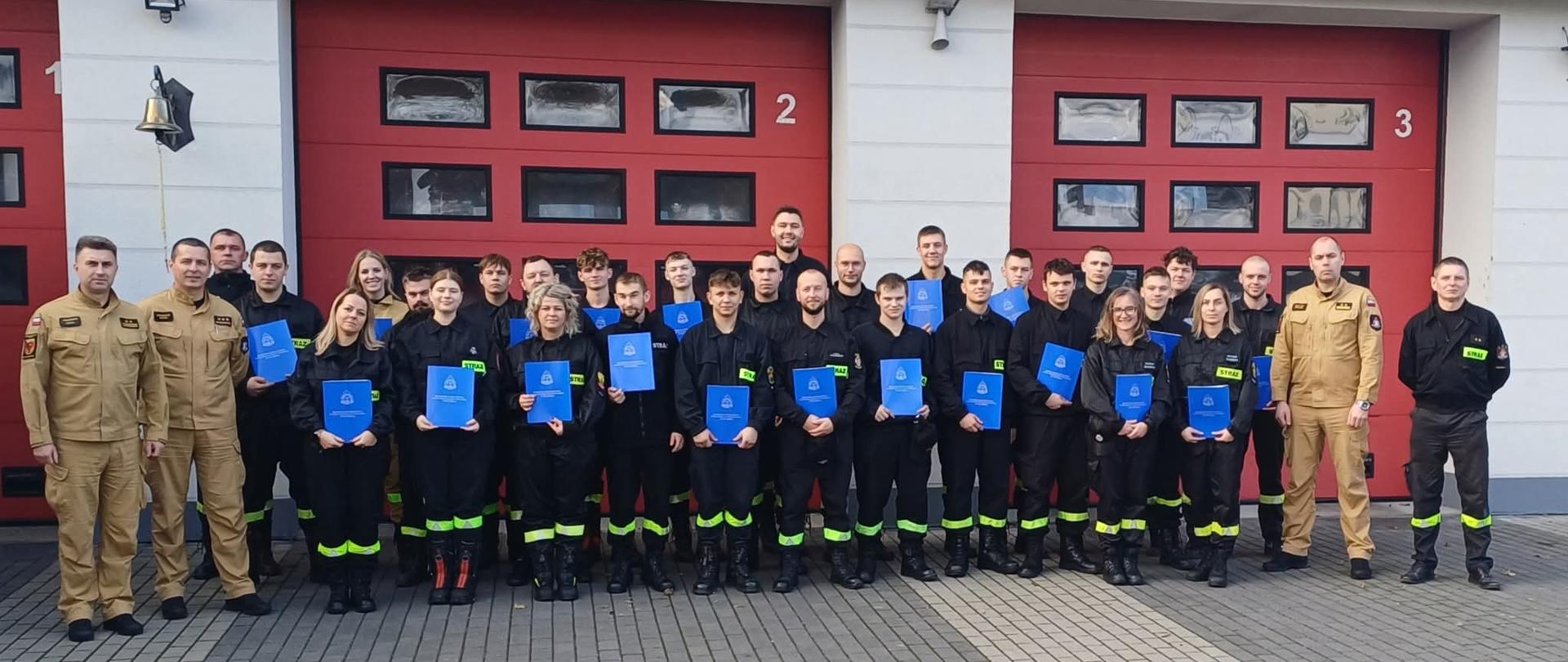 Grupa strażaków stoi na placu. Dwóch strażaków z lewej strony ma na sobie umundurowanie w kolorze żółtym. W środku strażacy mają umundurowanie w kolorze czarnym z elementami odblaskowymi. Z prawej strony stoi strażak w umundurowaniu koloru żółtego. Grupa strażaków w czarnym umundurowaniu trzyma w rękach niebieskie teczki. Za strażakami widać trzy czerwone bramy garażowe. Ściany budynku są koloru białego. 