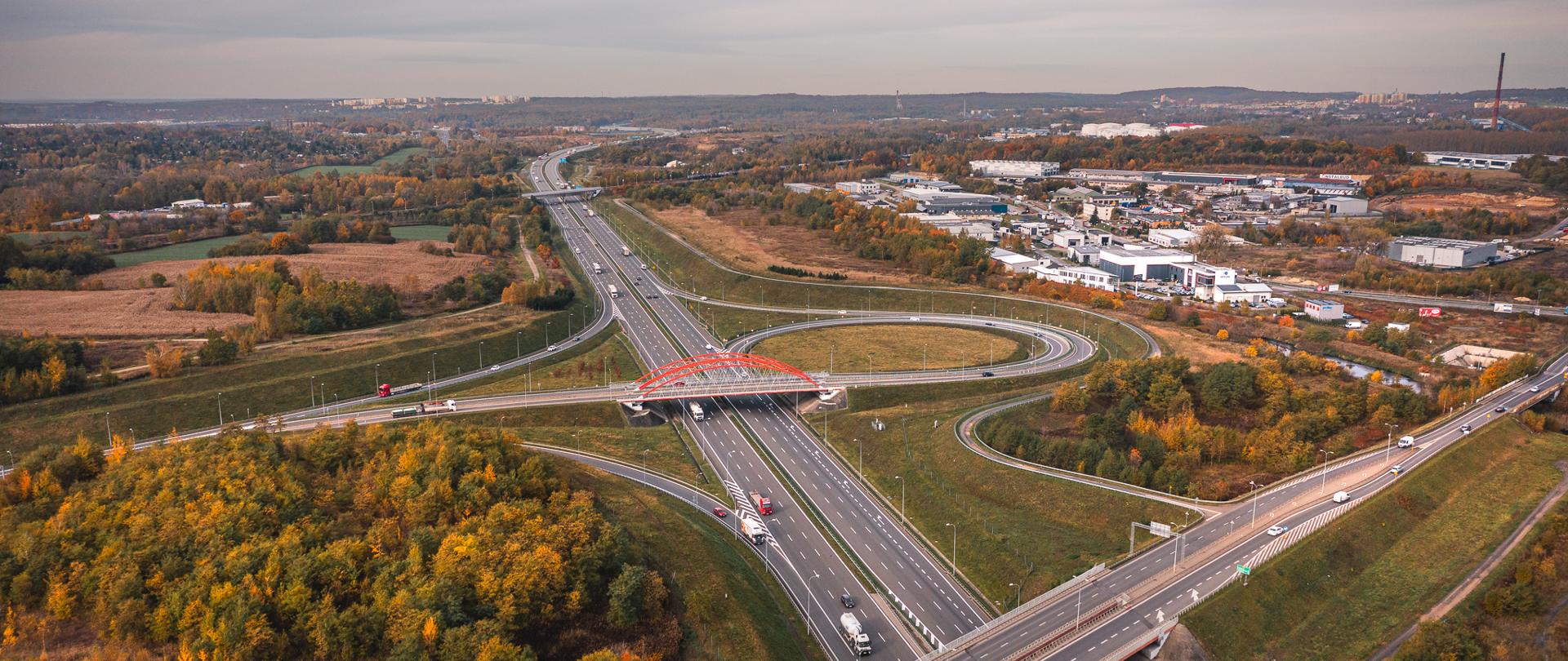 Węzeł drogowy harmonijnie wkomponowany w krajobraz miejski oraz częściowo leśny. Zielono-złociste barwy drzew wskazują, że zdjęcie wykonywane było jesienią. W prawym górnym rogu fotografii dostrzec możemy tereny przemysłowe oraz usługowo-handlowe.