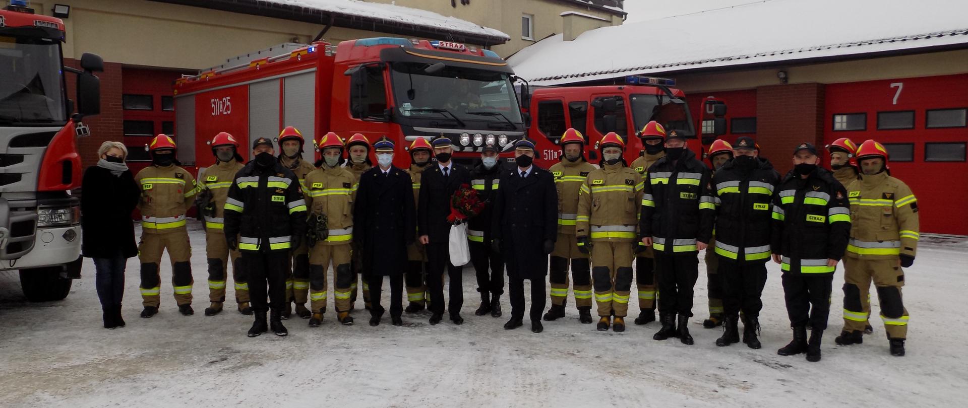 Dwudziestu dwóch strażaków i jeden pracownik cywilny stoją na placu. Pracownik cywilny – kobieta stoi po lewej stronie. Strażacy - większość w żółtych ubraniach specjalnych i hełmach. Cztery osoby w umundurowaniu dowódczo – sztabowym. Trzy osoby – Komendant, Jego zastępca i Tomasz Sokołowski w mundurach wyjściowych, stoją po środku na tle grupy. Wszyscy w maseczkach, Tomasz Sokołowski trzyma kwiaty i białą torebkę. W tle trzy samochody pożarnicze. Za nimi garaż z czerwonymi bramami. Wszyscy w maseczkach. Pora zimowa na dachu garażu i na placu śnieg.