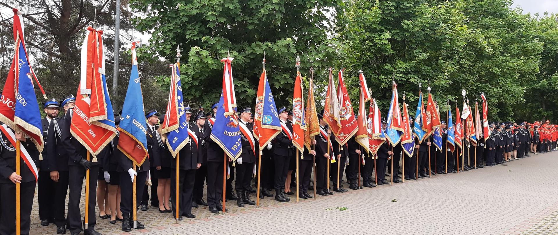 Na zdjęciu poczty sztandarowe jednostek Ochotniczych Straży Pożarnych powiatu pabianickiego, za nimi w po prawej pododdział OSP, a w tle w czerwonych koszulach stojące w szyku dzieci wchodzące w skład Młodzieżowej Drużyny Pożarniczej. 