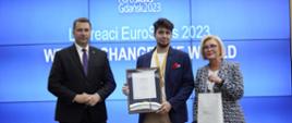 Minister Czarnek, wiceminister Machałek i młody człowiek w granatowej marynarce, z medalem na szyi stoją przed wielkim ekranem z napisem Laureaci EuroSkills 2023.