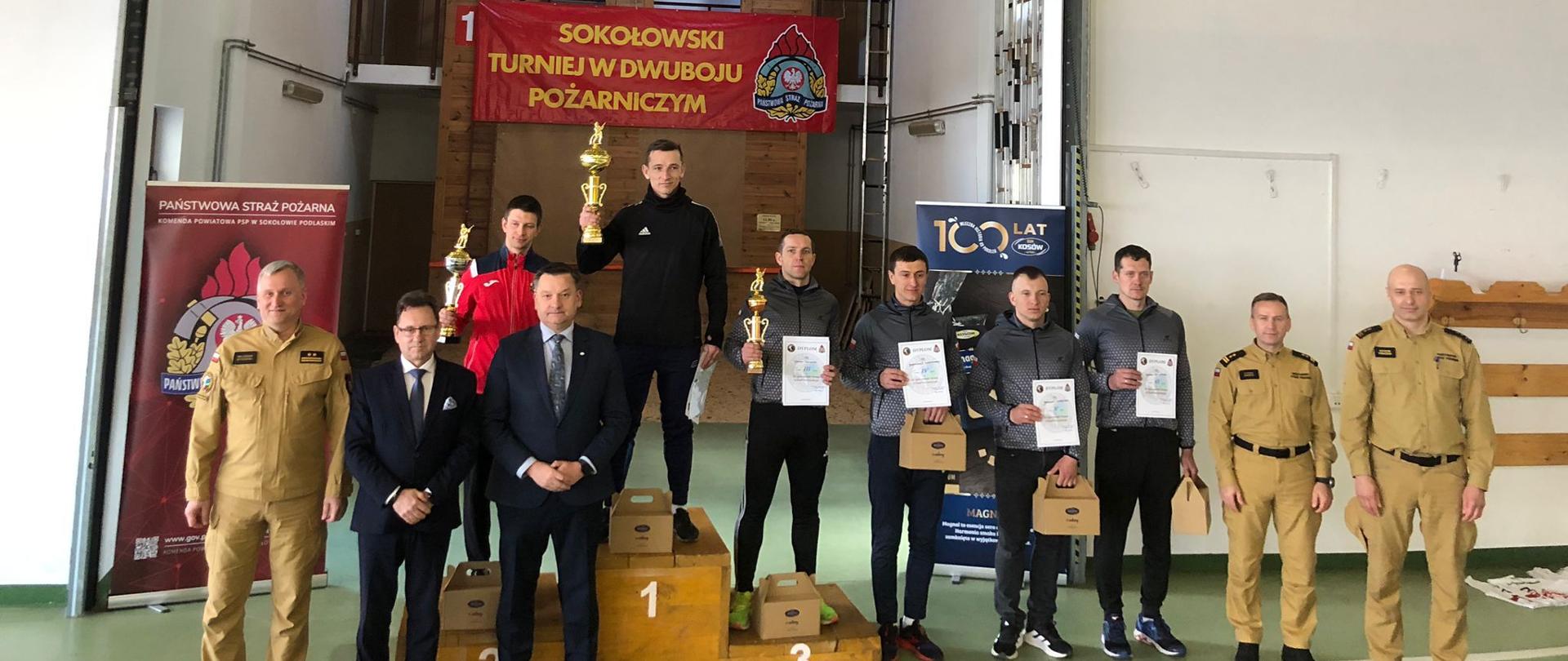Zdjęcie przedstawia zwycięzców zawodów w KP PSP Sokołów Podlaski z dyplomami i nagrodami rzeczowymi w rękach. Obok nich stoją funkcjonariusz PSP w ubraniach koszarowych koloru piaskowego i przedstawiciele władz powiatowych w garniturach. Zdjęcie wykonane na tle banerów KP PSP Sokołów Podlaski i OSM Kosów Lacki. Bardziej z tyłu widoczna wspinalnia.