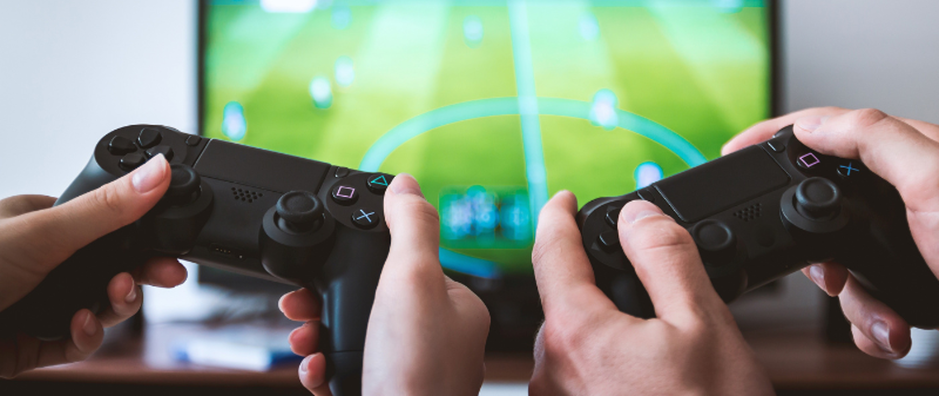 W dłoniach pady do gry wideo. W tle telewizor z wyświetlaną grą komputerową.