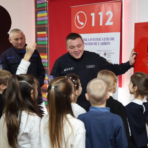 Strażak pokazuje dzieciom gaśnicę, grupka dzieci w wieku szkolnym go słucha, jedno dziecko podnosi rękę