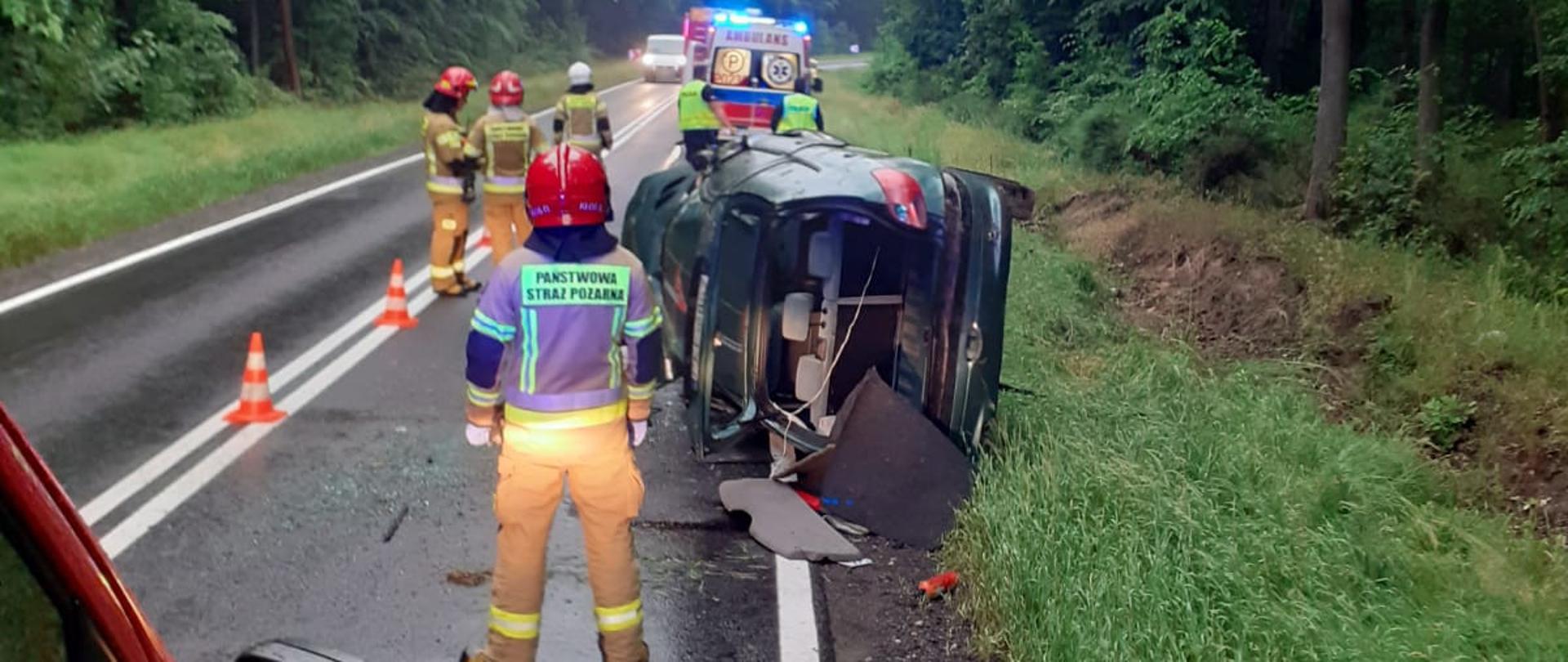 Widoczne miejsce zdarzenia: uszkodzony pojazd - samochód osobowy leżący na lewym boku; oraz służby działające na miejscu zdarzenia policja, pogotowie ratunkowe i straż pożarna