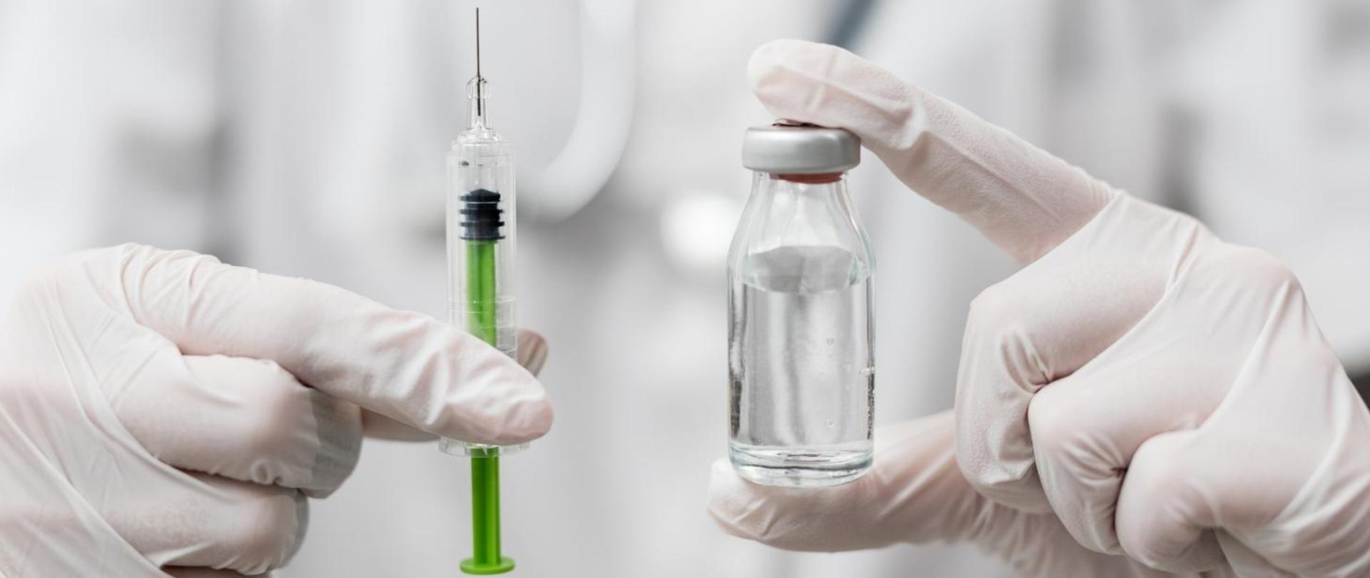 Zdjęcie przedstawia ręce w ochronnych rękawiczkach trzymających w jednej strzykawkę a w drugiej ręce fiolkę z przezroczystym płynem.