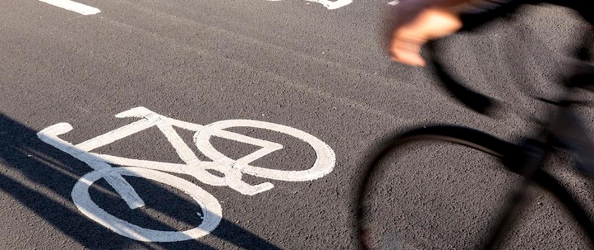 Osoba dojeżdżająca do pracy na ścieżce rowerowej. Na czarnej jezdni narysowany biały rower. Na pierwszym planie z prawej strony widok koła rowerowego i rąk na kierownicy.