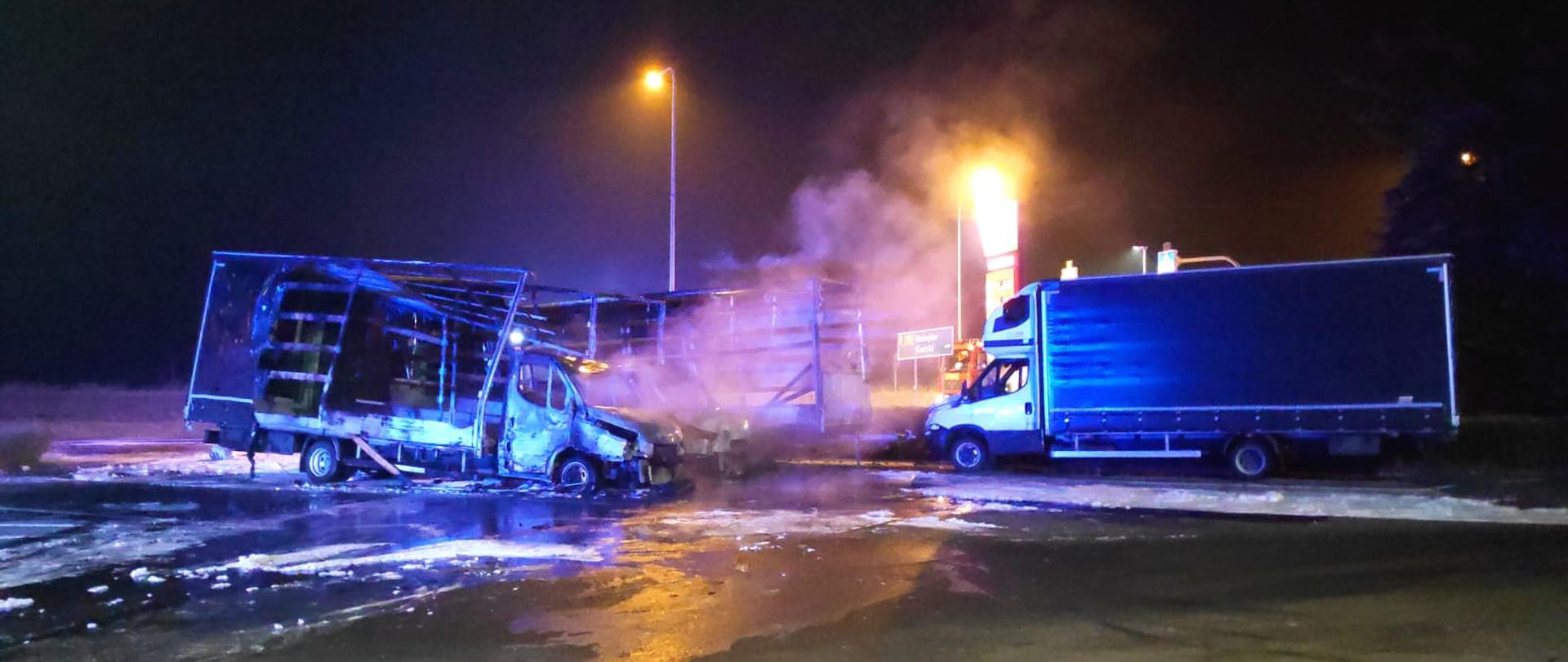 Na zdjęciu dwa spalone samochody dostawcze oraz jeden samochód uratowany. W tle stacja benzynowa.