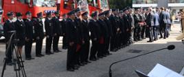 Strażacy ochotniczych straży pożarnych ustawieni w dwóch rzędach otrzymują czerwone teczki wręczane przez strażaków Państwowej Straży Pożarnej wojewodę pomorskiego oraz posłów obok stoi statyw za nimi stoją samochody pożarnicze.