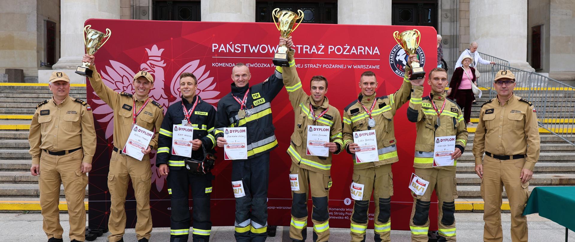 Laureaci kategorii mężczyzn pozują do wspólnego zdjęcia podczas ceremonii dekoracji przed Pałacem Kultury i Nauki w Warszawie. Zawodnicy prezentują puchary, medale i dyplomy zdobyte w 8. Mistrzostwach Polski Strażaków w Biegu po Schodach „Wieżowiec 2026”, w towarzystwie przedstawicieli Państwowej Straży Pożarnej.