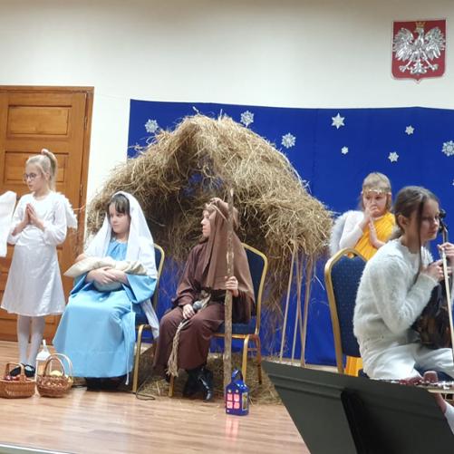 Na scenie uczniowie przebrani za Maryję i Józefa siedzą na krzesłach a w tle szopka. 