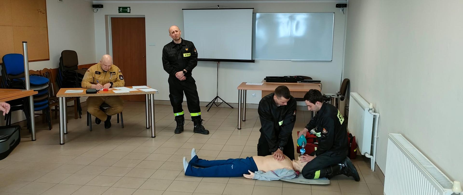 W sali edukacyjnej Komendy Powiatowej Państwowej Straży Pożarnej w Rypinie odbywa się egzamin recertyfikujący uprawnienia z kwalifikowanej pierwszej pomocy. Z prawej strony dwoje funkcjonariuszy udziela kwalifikowanej pierwszej pomocy fantomowi, który imituje osobę poszkodowaną. Ich pracę nadzorują młodszy brygadier Marek Ruciński wojewódzki koordynator ratownictwa medycznego Państwowej Straży Pożarnej województwa kujawsko-pomorskiego oraz starszy ogniomistrz Mirosław Błażejczyk koordynator powiatowy ratownictwa medycznego Państwowej Straży Pożarnej.