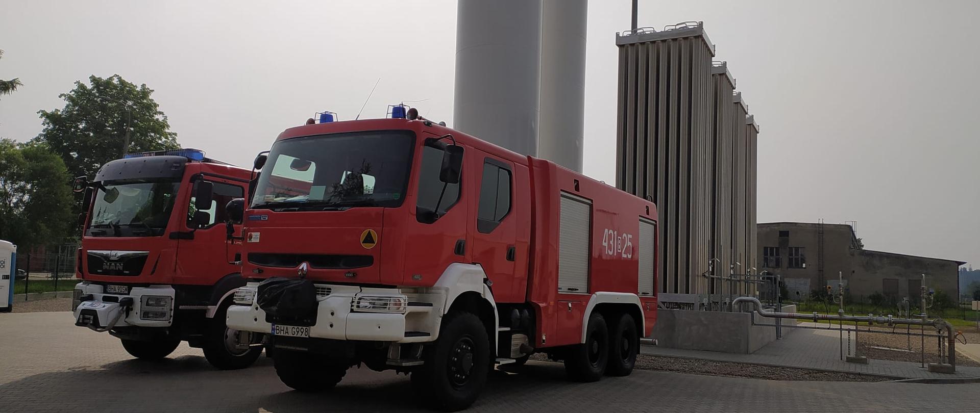 Dwa strażackie samochody stoją na tle zbiorników z gazem LNG.