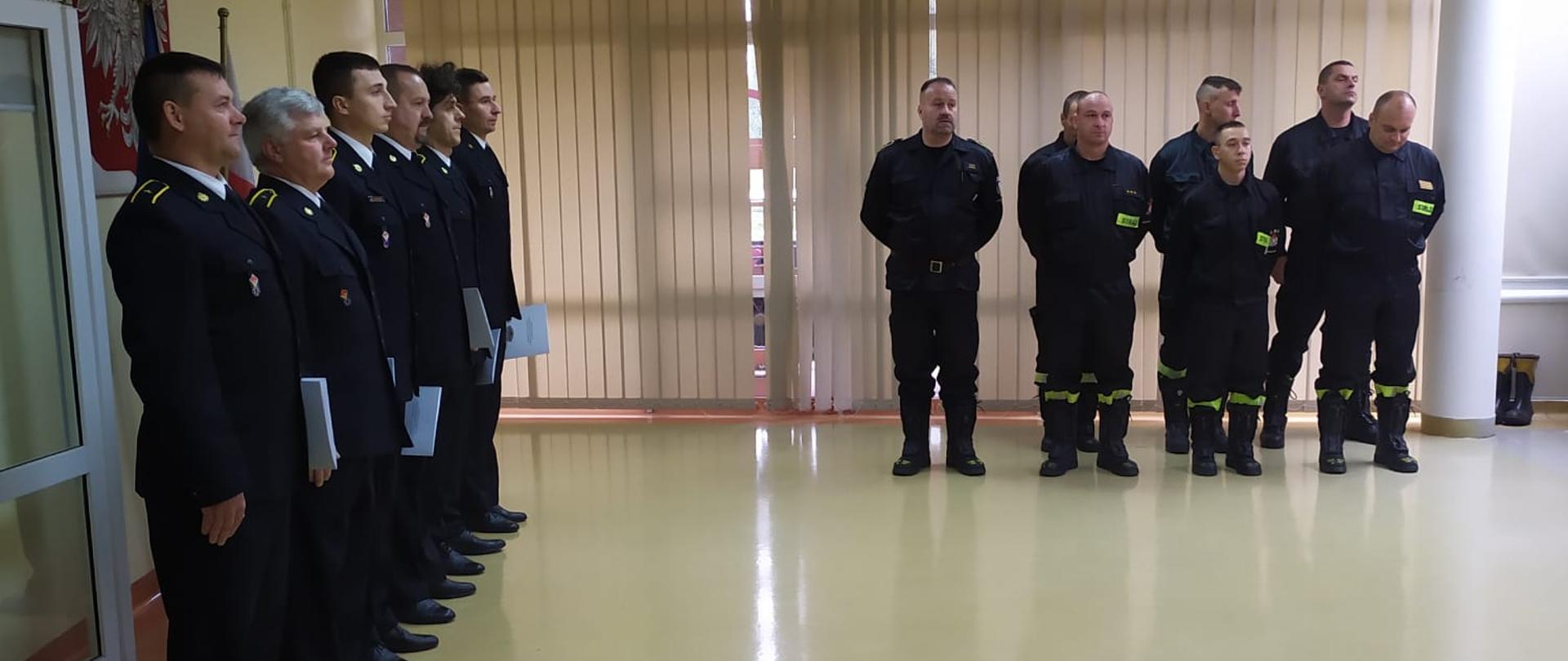Uroczystość wręczenia awansów w Sali Sztabowej Komendy Powiatowej Państwowej Straży Pożarnej w Kamiennej Górze. Jeden szereg strażaków stoi po prawej stronie drugi szereg na wprost oglądającego zdjęcie. Funkcjonariusze zwróceni twarzami do przemawiającego. Przemawiający niewidoczny na zdjęciu.