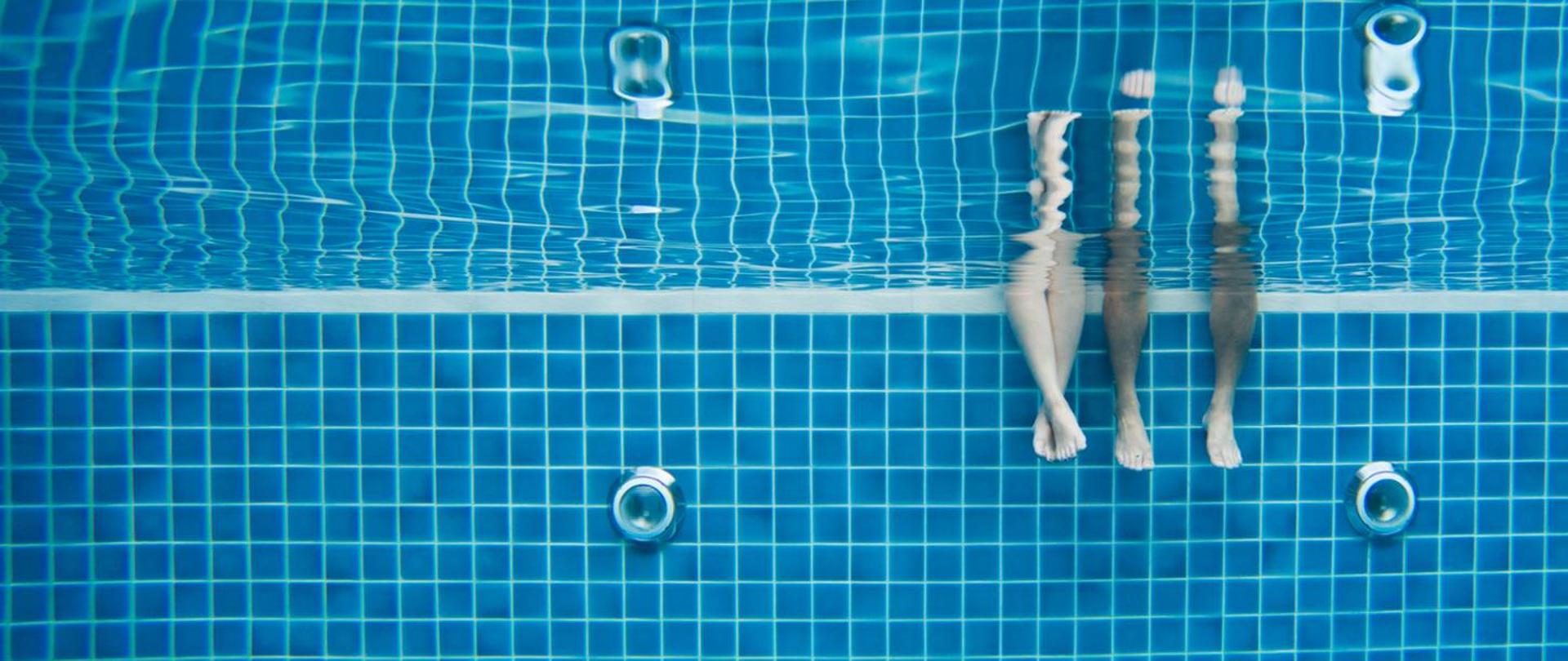 Podwodne zdjęcie basenu. W prawej części zdjęcia dwie pary nóg. Ze skrajnej strony męskie nogi, obok z lewej strony damskie.