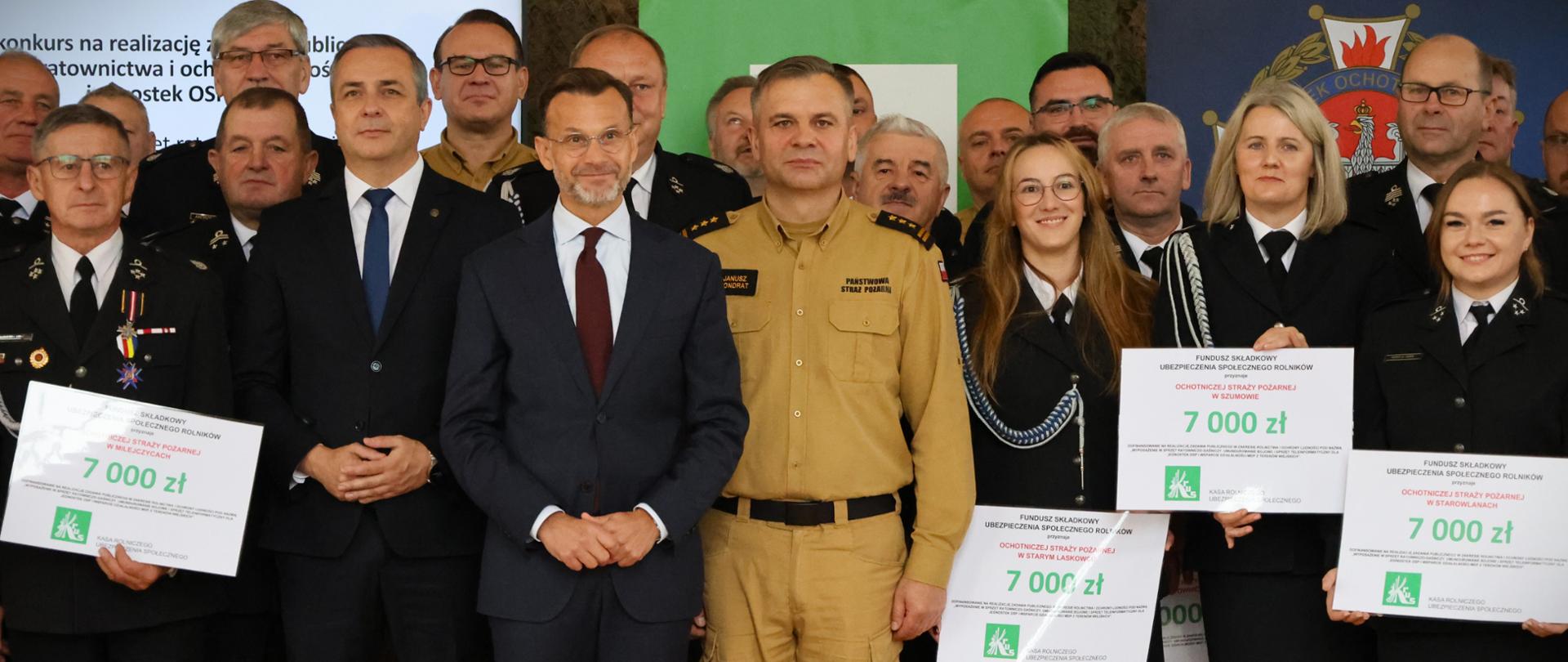 Wsparcie finansowe przekazane przez Kasę Rolniczego Ubezpieczenia społecznego na rzecz 52 jednostek OSP z terenu województwa podlaskiego - zebrani goście w towarzystwie organizatorów przedsięwzięcia i druhów ochotników, niektórzy trzymają bony