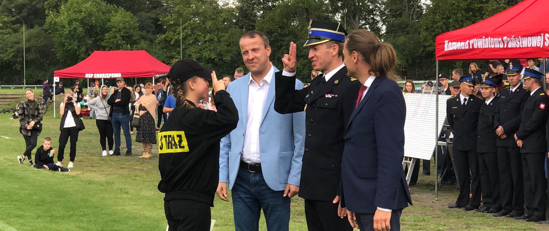 salutująca dziewczynka w mundurze strażaka, przed nią stoi trzech mężczyzn . w srodku salutrujący strażak , w tle grupa sstrażaków stojących pod czerwonym namiotem