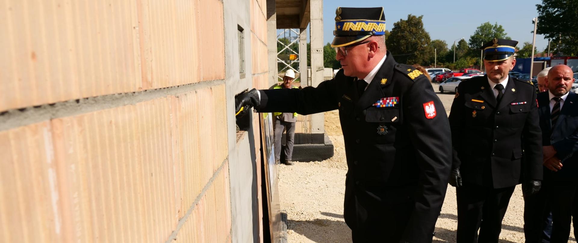 Zastępca komendanta głównego PSP na małej łopatce wkłada trochę zaprawy budowlanej na mur nowej komendy