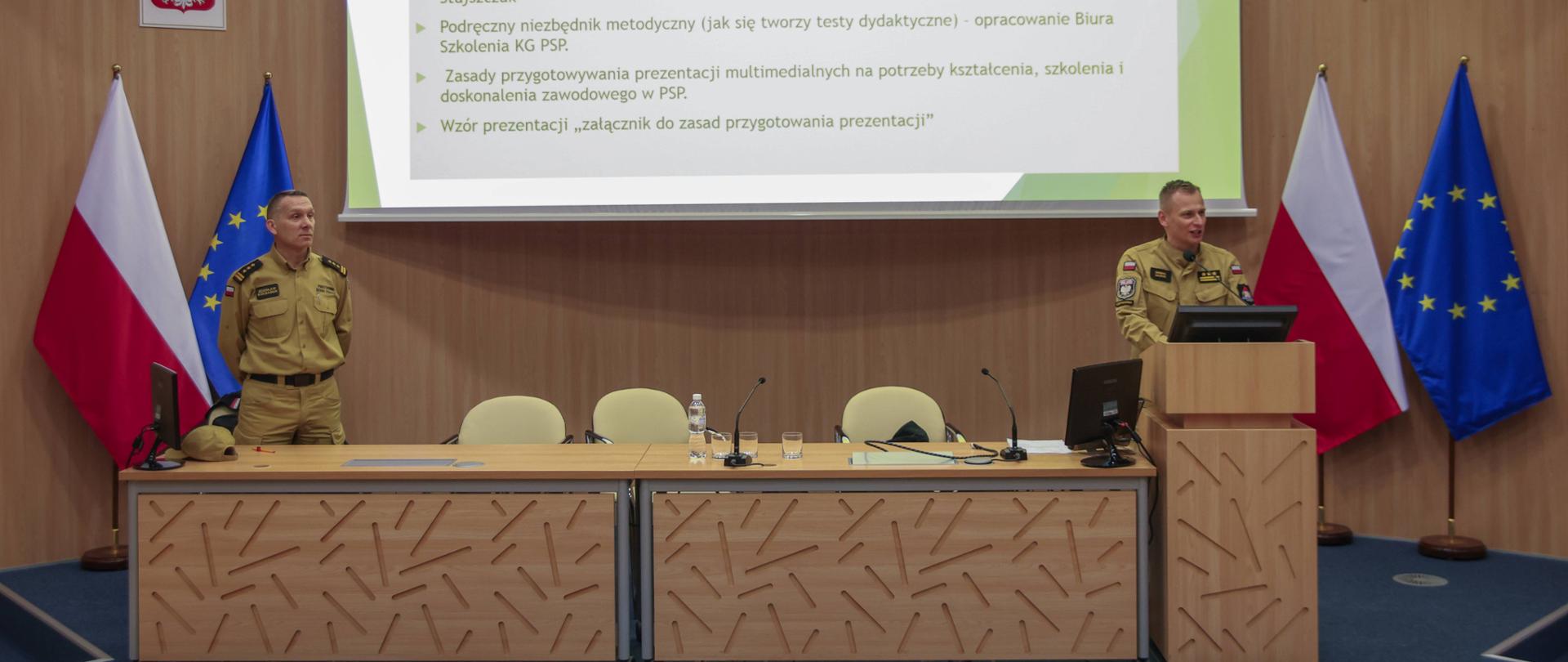 Po lewej strażak stojący za stołem konferencyjnym, po prawej Zastępca Komendanta Centralnej Szkoły PSP przemawiający stojąc na mównicy, w tle ekran multimedialny z wyświetlonym slajdem prezentacji
