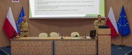 Po lewej strażak stojący za stołem konferencyjnym, po prawej Zastępca Komendanta Centralnej Szkoły PSP przemawiający stojąc na mównicy, w tle ekran multimedialny z wyświetlonym slajdem prezentacji