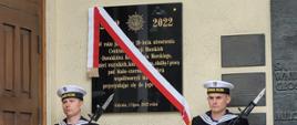 Żołnierze Marynarki Wojennej stoją z pistoletami obok tablicy pamiątkowej z biało-czerwoną wstęgą.