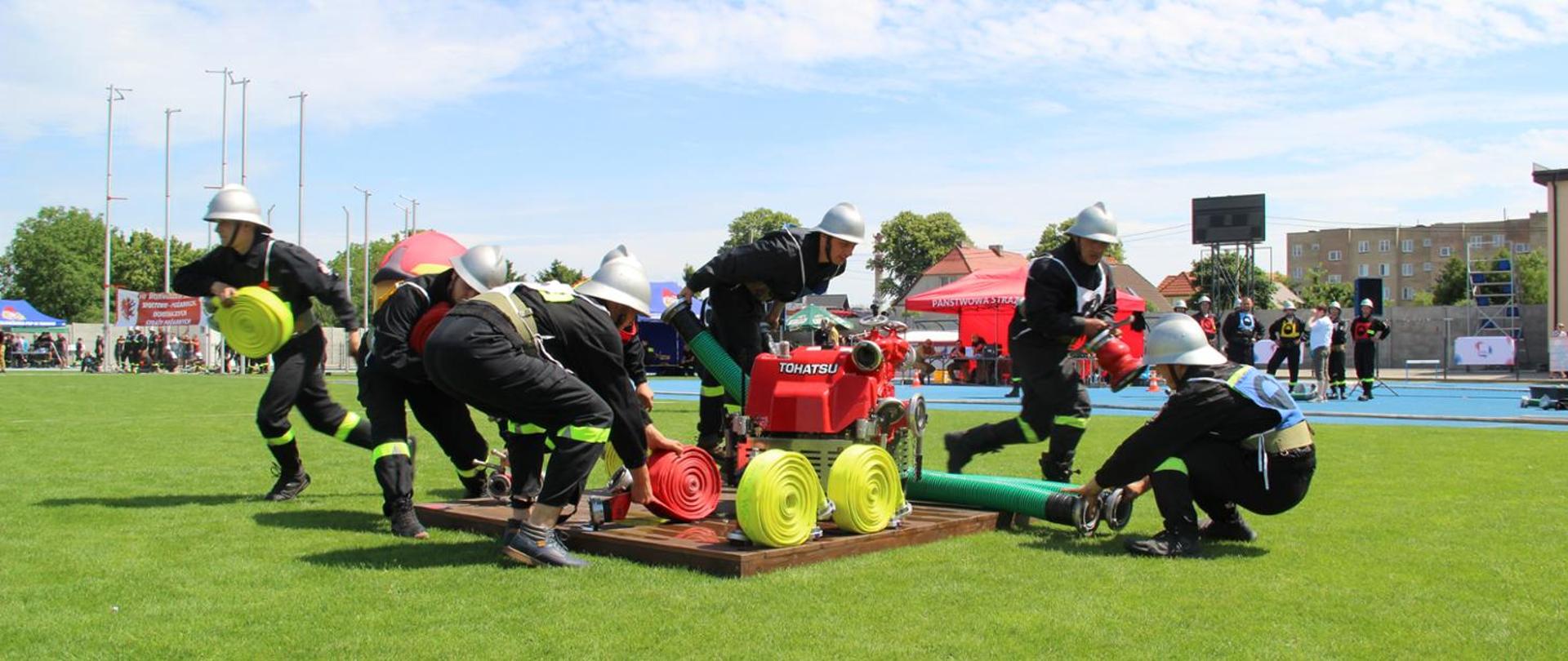 Na podeście motopompa strażacka. Wokół niej zawodnicy - strażacy w strojach sportowych, podejmują węże i sprzęt. W tle dmuchany hełm strażacki, reklamowy. Dalej obserwatorzy