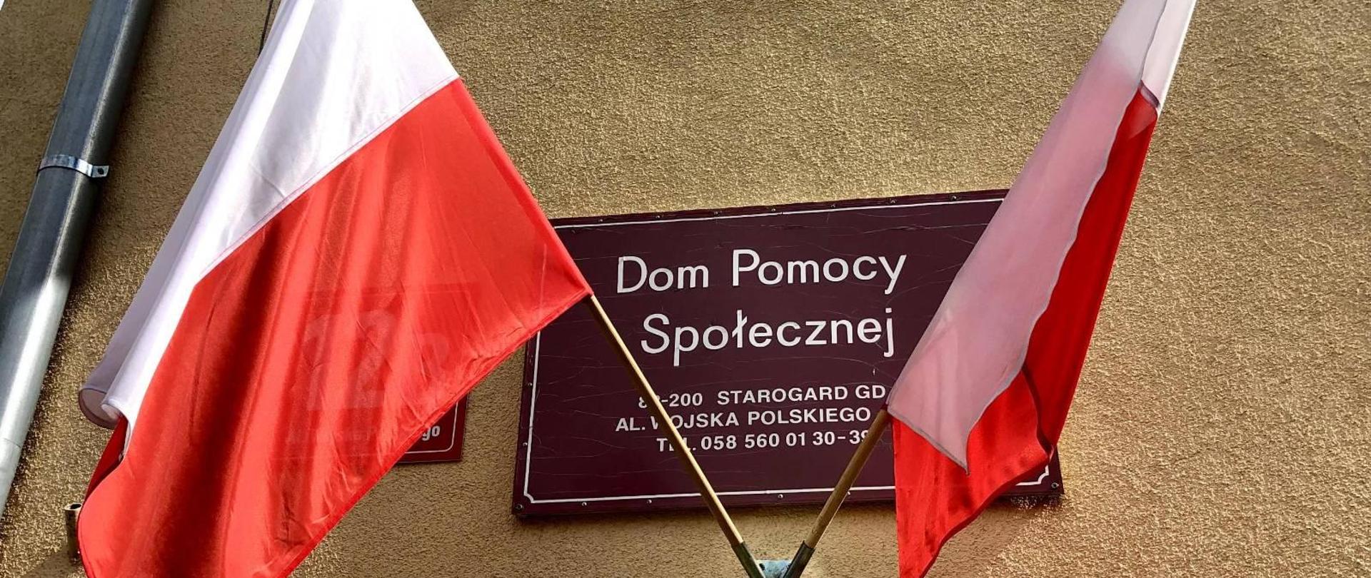 Zdjęcie przedstawia tablicę z napisem Dom Pomocy Społecznej, poniżej dwie flagi państwowe 
