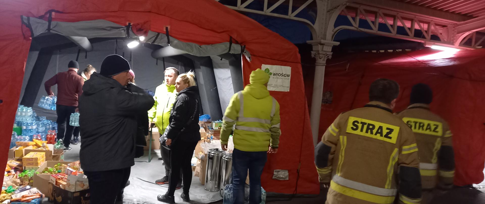 Widoczne osoby cywilne i umundurowanie strażacy przed namiotami pneumatycznymi ustawionymi pod zadaszeniem dworca koleinowego. W pierwszym od lewej namiocie widoczne wewnątrz ustawione produkty żywieniowe i napoje.