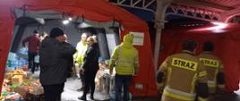 Widoczne osoby cywilne i umundurowanie strażacy przed namiotami pneumatycznymi ustawionymi pod zadaszeniem dworca koleinowego. W pierwszym od lewej namiocie widoczne wewnątrz ustawione produkty żywieniowe i napoje.