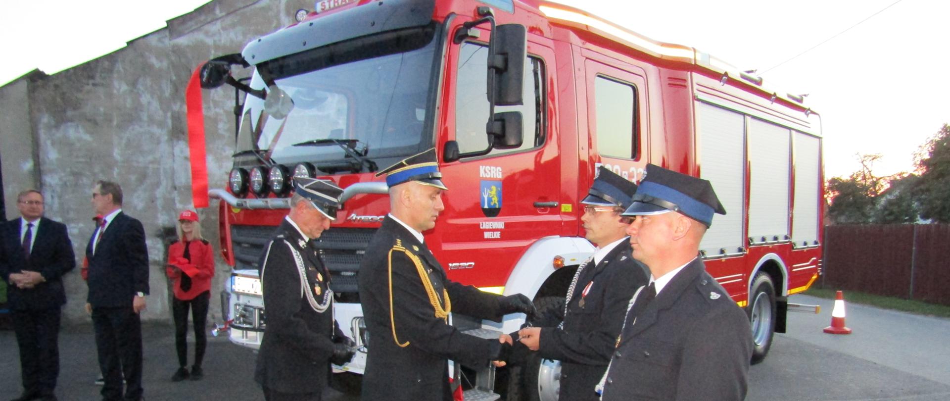 Przed pojazdem cztery osoby, jedna z nich przekazuje kluczyki samochodowe. W tle samochód pożarniczy.