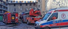 Dwa samochody strażackie i karetka pogotowia stojące przy budynku mieszkalnym