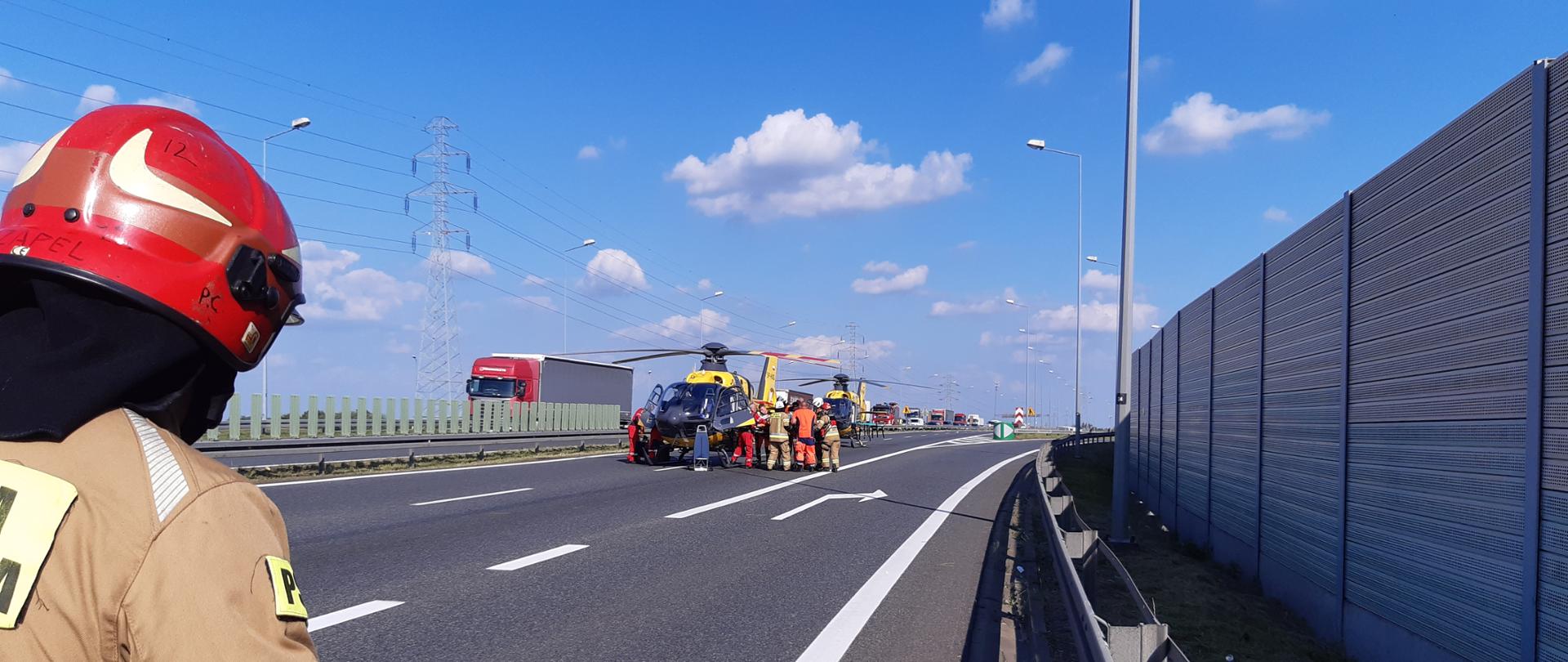 Na zdjęci po lewej stronie widoczny strażak stojący strażak w hełmie i ubraniu specjalnym patrzący na znajdujący się w oddali śmigłowiec LPR