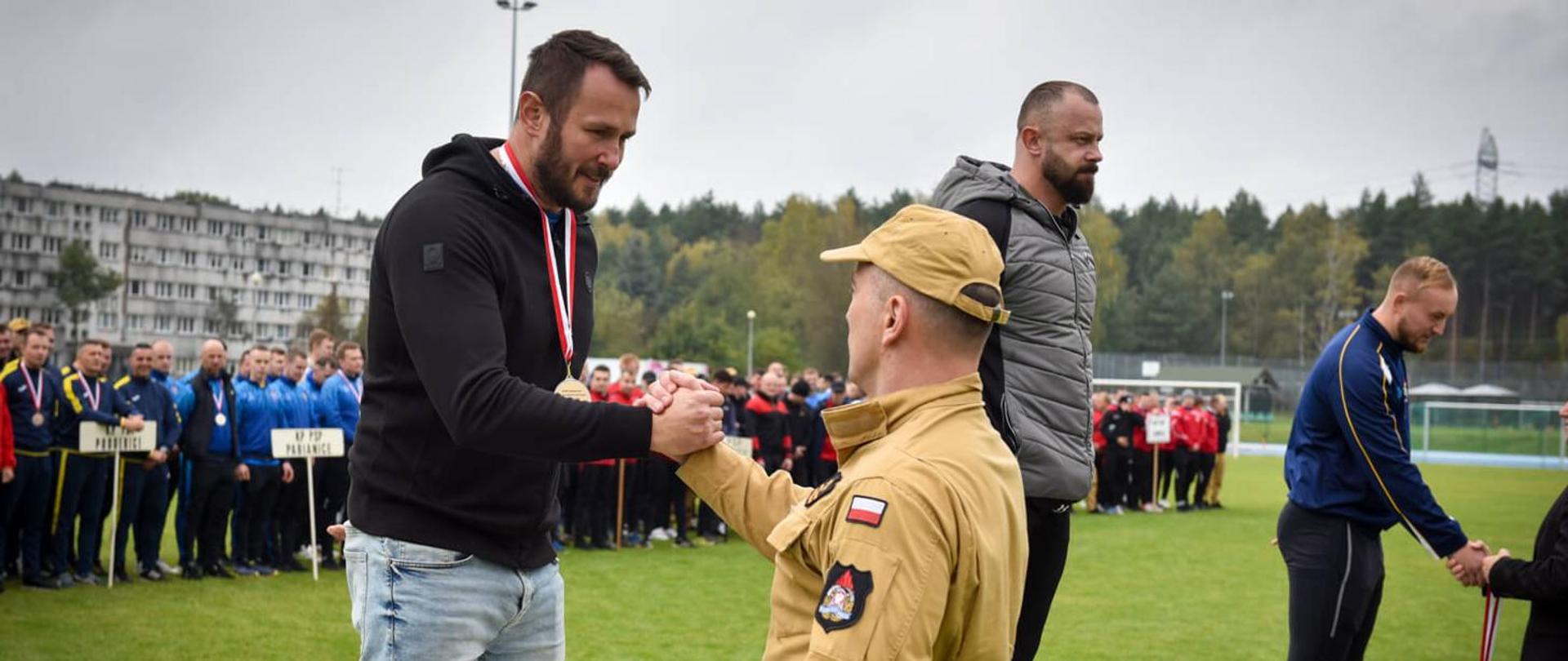 Mężczyzna w czarnej bluzie i jeansach, ze medalem na szyi, ściska dłoń mężczyzny w beżowym mundurze na podium, podczas ceremonii wręczenia nagród