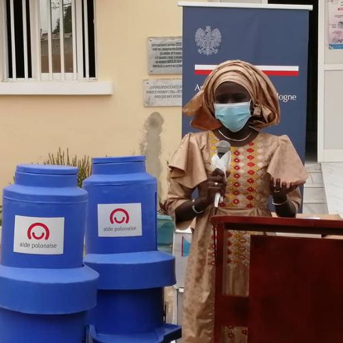 kobieta w tradycyjnym stroju trzyma mikrofon, przy niej stoją niebieskie pojemniki na wodę