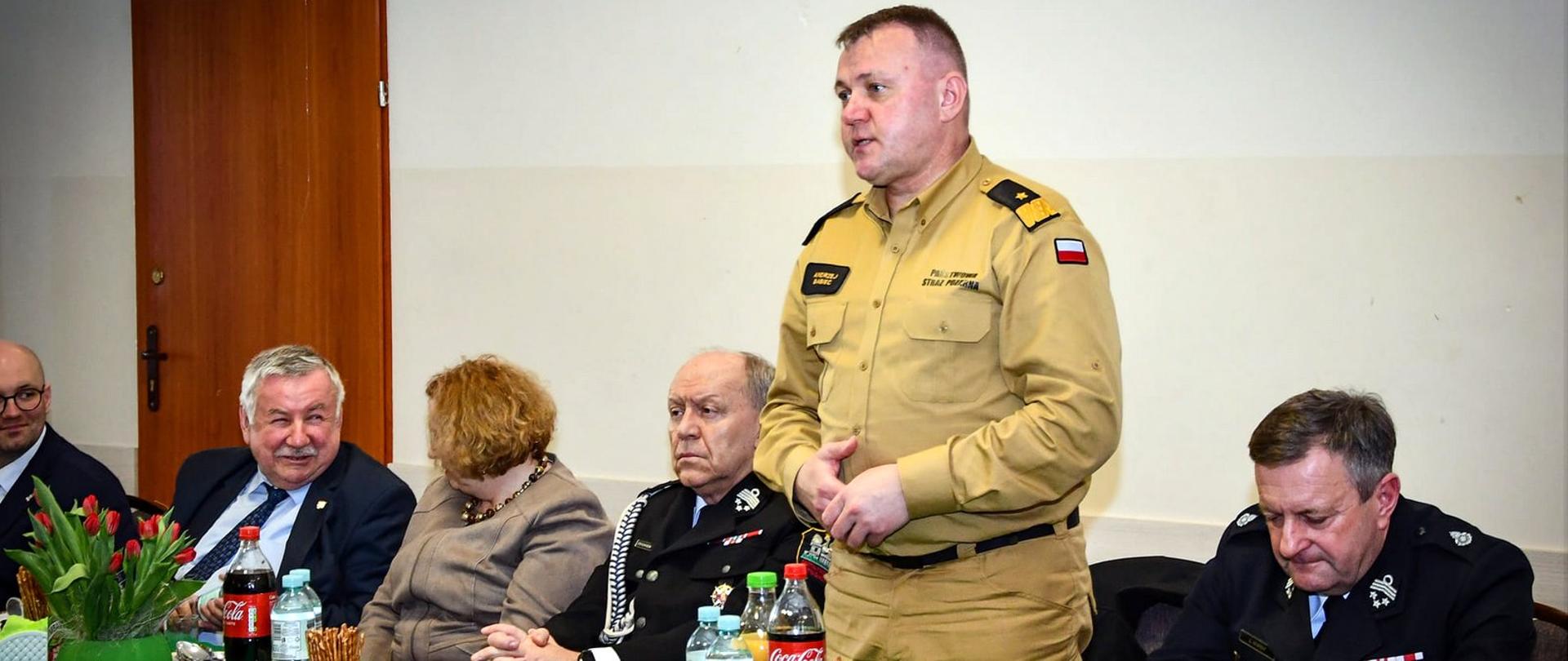 Zdjęcie zrobione w pomieszczeniu. Za stołami siedzą uczestnicy spotkania z druhami ochotnikami. Wśród nich strażacy ochotnicy oraz strażacy Państwowej Straży Pożarnej. Oficer Państwowej Straży Pożarnej w stopniu generała stoi i przemawia.