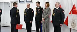 Funkcjonariusz Państwowej Straży Pożarnej w mundurze wyjściowym stoi na baczność trzymając czerwoną teczkę przed nim stoi komendant główny Państwowej Straży Pożarnej wojewoda pomorska oraz kadra kierownicza Państwowej Straży Pożarnej z terenu województwa pomorskiego za nimi stoją trzy flagi Polski na ścianie wisi rota ślubowania strażackiego.