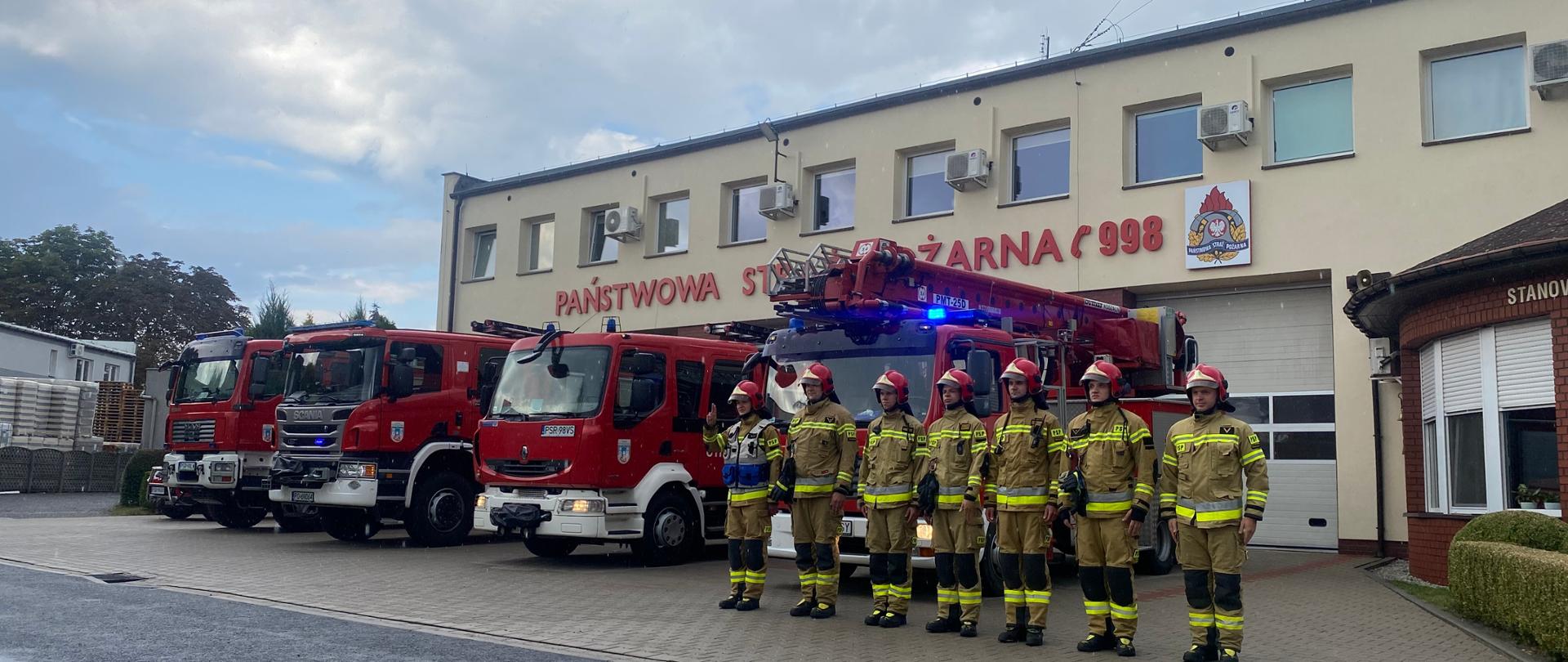 Strażacy JRG Środa Wielkopolska podczas uroczystej zbiórki z okazji 78 Rocznicy Powstania Warszawskiego. Strażacy w szeregu w umundurowaniu bojowym na placu przed budynkiem KP PSP Środa Wielkopolska. W tle samochody pożarnicze z włączonymi sygnałami świetlnymi. Dowódca pierwszy po lewej salutuje.