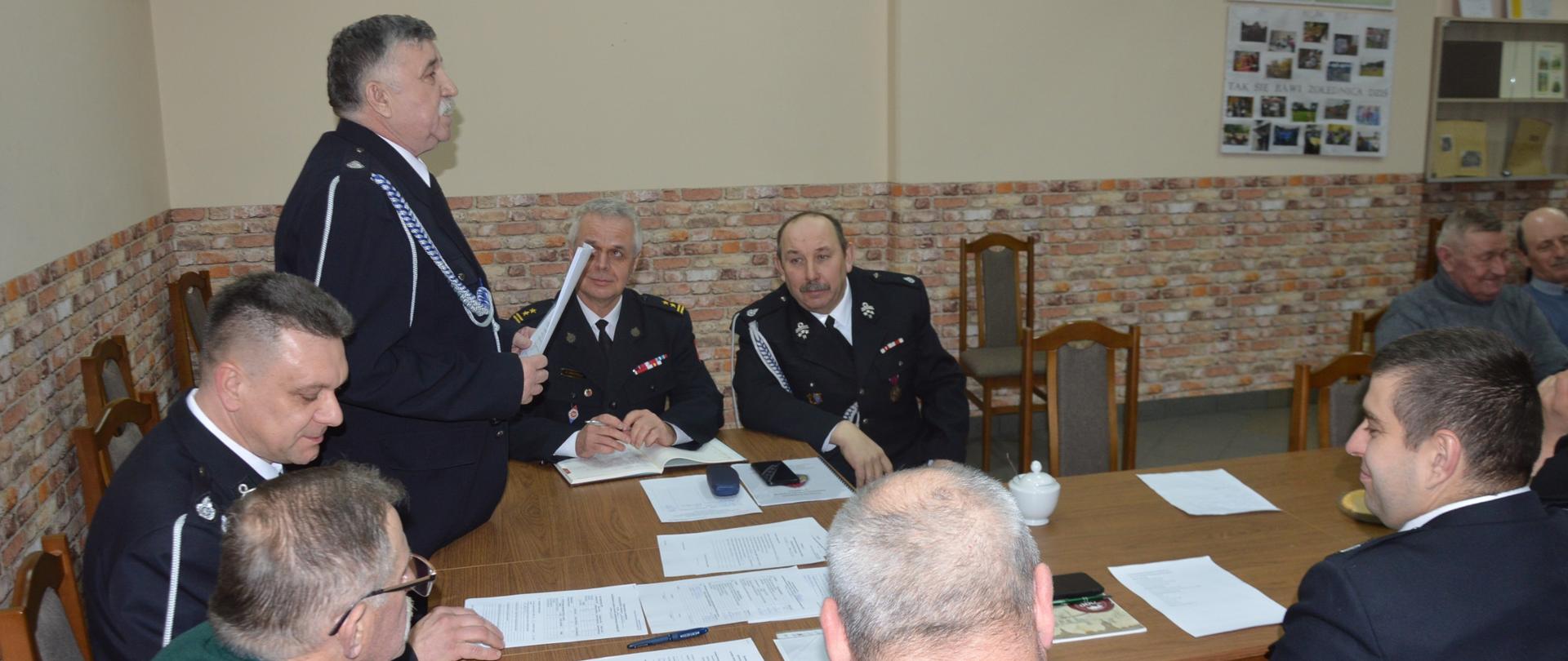 Zebranie sprawozdawcze OSP Żołędnica. Świetlica wiejska w Żołędnicy. Za stołem prezydialnym siedzą zaproszeni goście oraz przedstawiciele zarządu miejscowej OSP. Przewodniczący zebrania na stojąco prowadzi obrady. Na stole leżą dokumenty.