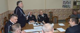 Zebranie sprawozdawcze OSP Żołędnica. Świetlica wiejska w Żołędnicy. Za stołem prezydialnym siedzą zaproszeni goście oraz przedstawiciele zarządu miejscowej OSP. Przewodniczący zebrania na stojąco prowadzi obrady. Na stole leżą dokumenty.