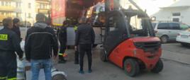 Sprzęt pożarniczy dla ukraińskich strażaków. Komenda Powiatowa PSP w Gostyniu. Strażacy przygotowują do transportu zebrany sprzęt. Na placu komendy stoi samochód ciężarowy, na który za pomocą wózka widłowego ładowana jest europaleta ze sprzętem. Obok samochodu znajduje się 6 strażaków. W tle budynek mieszkalny wielorodzinny. 