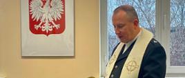 Kapelan w stroju liturgicznym czytający modlitwę podczas uroczystości, stojący przy nakrytym stole z poczęstunkiem. W tle widoczne jest godło Polski oraz okno z widokiem na drzewa.