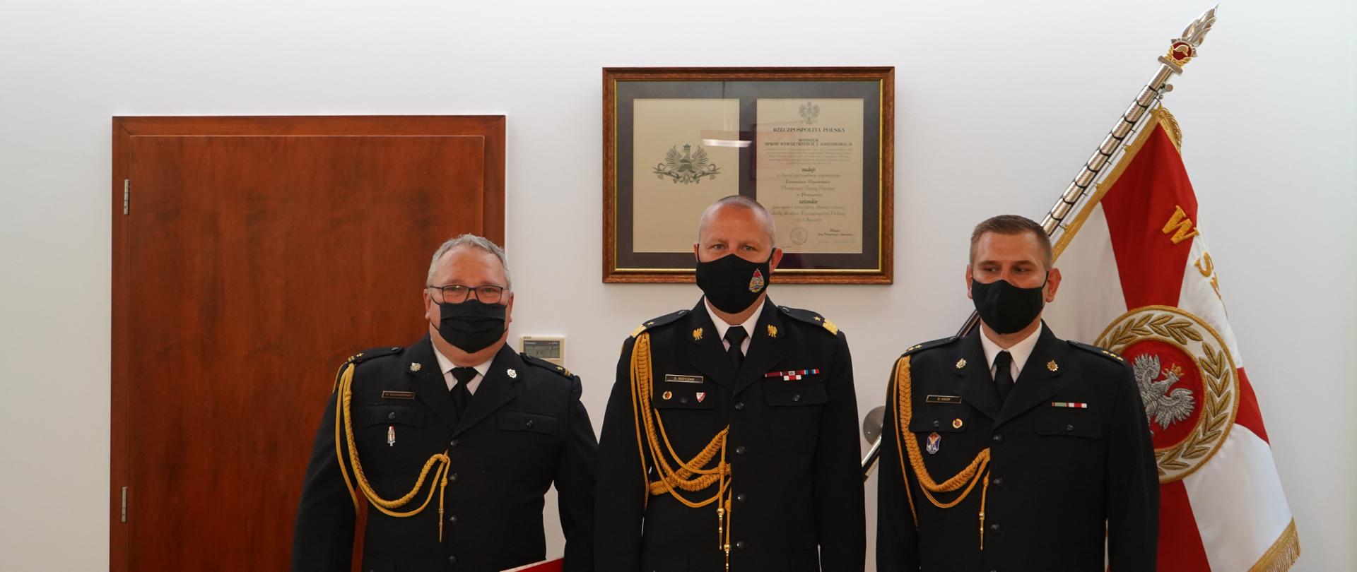 trzech strażaków w ciemnych mundurach stoi na tle sztandaru