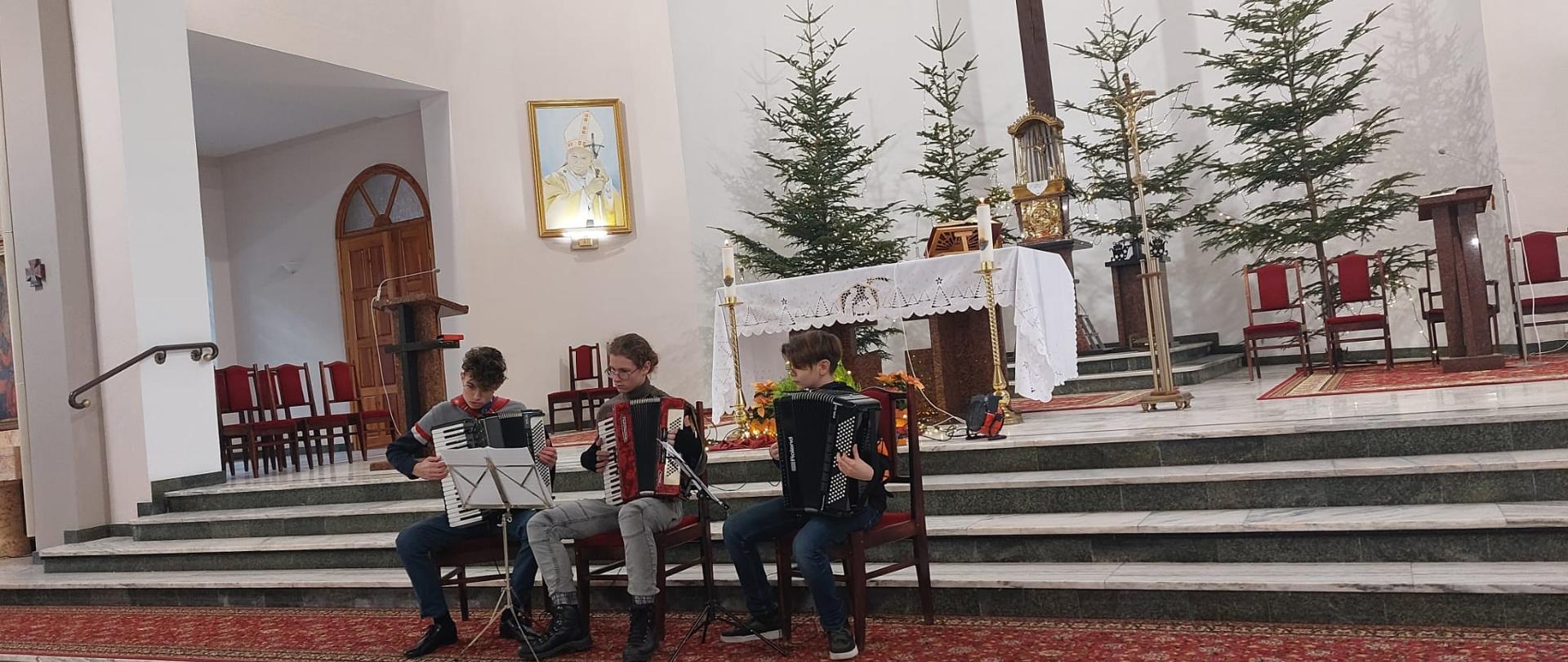 Zdjęcie trójki uczniów z klas akordeonu grających na akordeonie, Zdjęcie wykonane w Kościele Podwyższenia Krzyża w Kłodzku, w tle widoczny ołtarz kościoła.