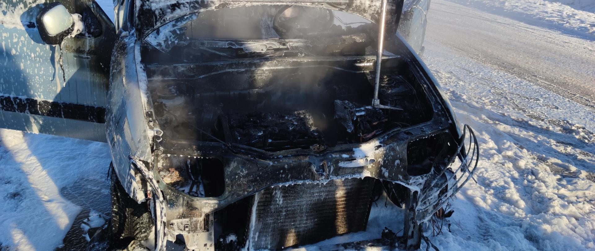 Zdjęcie przedstawia samochód z otwartą maską stojący na zaśnieżonej ulicy. W środku widać spaloną komorę silnika oraz lekki dym