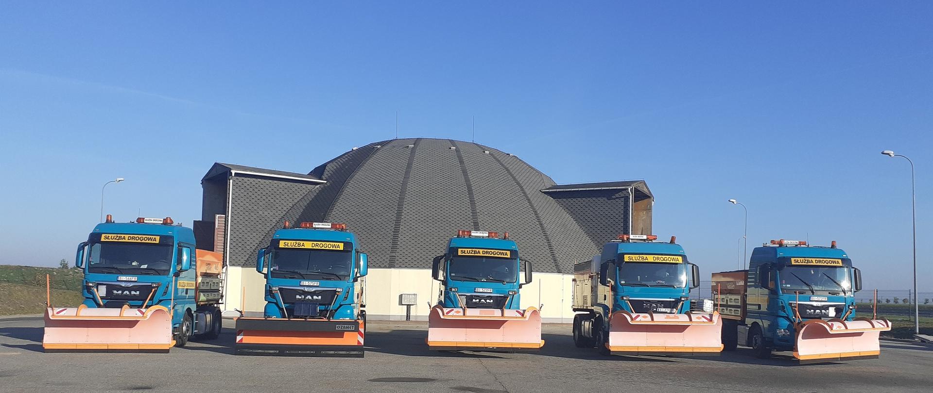 Pięć pojazdów ciężarowych z zamontowanymi pługami odśnieżnymi na tle magazynu soli.