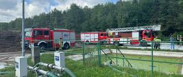 Trzy samochody pożarnicze, strażacy, oraz pracownicy firmy na terenie składowiska przy zbiorniku wodnym.
